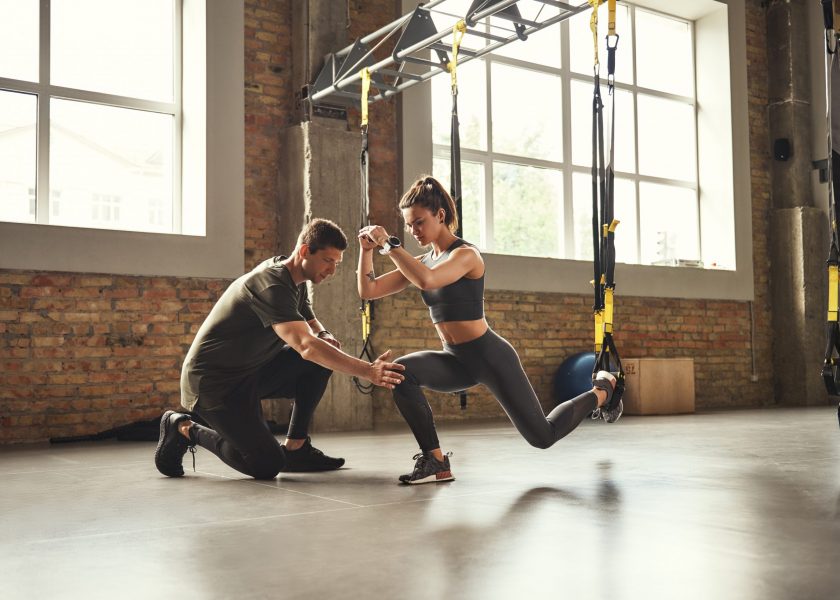 Doing squat exercise. Confident young personal trainer is showing slim athletic woman how to do squats with Trx fitness straps while training at gym.