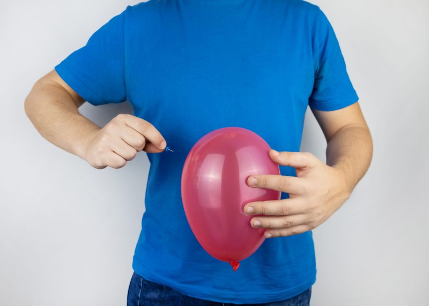 Conceptual photography. The man holds a red ball near his belly, which symbolizes bloating and flatulence. Then he brings a needle to it to burst the balloon and thus get rid of the problem.